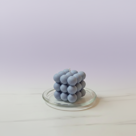 A blue bubble-shaped soy wax candle sits on a clear glass dish against a light purple background.